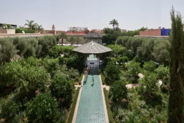 A view into a walled garden. There is a green tiled pathway and a covered seating area surrounded by various green trees.