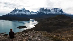 A female hiker is sitting on rocks and looking out across a turquoise lake and snow-capped mountains in Torres del Paine National Park, Chile.