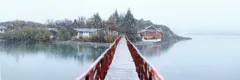 Snow is falling on a red wooden bridge, it crosses the water to a small, tree covered island, where the red buildings of Hosteria Pehoe can be seen.