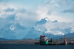 Two boats are docked outside of Punto Notales, Chile. Snow-capped mountains partly covered by clouds are in the background.