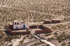 A small white church is ringed by a red stone wall. It is surrounded by desert and shrubs.