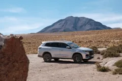 A silver car is parked in the Atacama Desert, surrounded by grass, shrubs and a mountain.