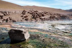 Pools of turquoise water are steaming, surrounded by red rock at Geyser Blanco in the Atacama Desert, Chile.