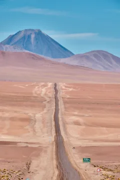 A long road runs through the Atacama Desert with hills and volcanoes in the distance.