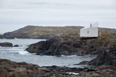 A single white modern building sits on a rocky outcrop, overlooking the ocean.