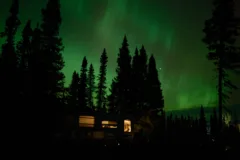 An RV sits in the dark, the lights are on inside. Outside, the silhouettes of trees are backed by the glowing green of the Northern Lights.