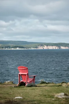 A red Adirondack chair sits on a grassy cliff-edge overlooking the ocean.