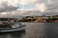 A white boat floats in the water. There is a collection of small white houses on top of the rocky cliffs behind it.