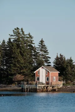 A red wooden boat house sits on the waters edge and is backed by trees.