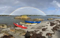 3 colourful kayaks sit coming out of the ocean onto a rocky beach. There are islands in the distance and a rainbow frames them.