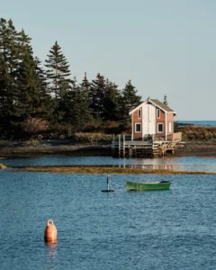 A bouy and fishing boat float in the foreground. A wooden boat house is backed by trees behind them.