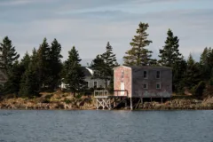 An old wooden boathouse sits in front of a small house. They are backed by trees.