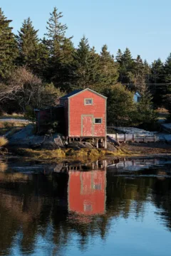 A red boat house on stilts sits raised out of the ocean. It's backed by trees.