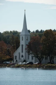 A white church with a tall, narrow steeple, sits on the waters edge. It's surrounded by trees.