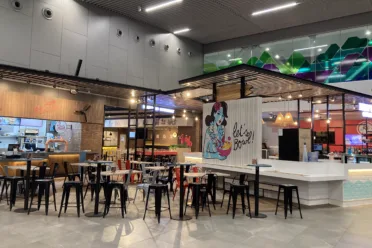 Metal chairs and tables surrounded by restaurants in a food court. There is a mural of a women holding a poke bowl on the wall.