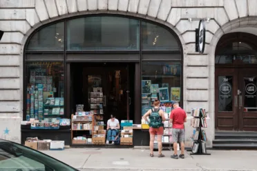 A young woman sits reading on the stairs of a bookshop. A couple stand browsing the book stands on the street outside.