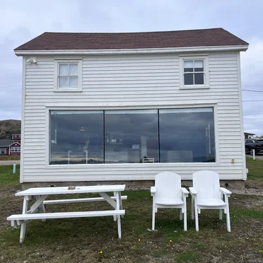 A white wooden house with a large picture window downstairs. there's a bench and chairs in the garden.