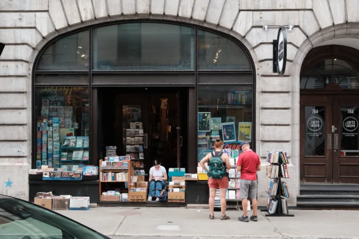 A young woman sits reading on the stairs of a bookshop. A couple stand browsing the book stands on the street outside.