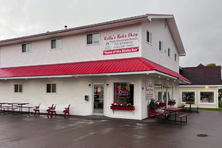 A large white building with a red roof. The sign on the side reads "Kelly's Bakery". There is an open sign in the window.