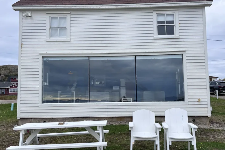 A white wooden house with a large picture window downstairs. there's a bench and chairs in the garden.