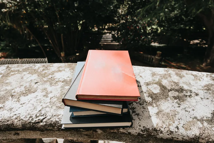 A stack of old, colourful books on a stone balcony that over looks a park.