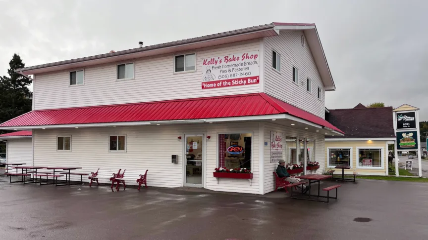 A large white building with a red roof. The sign on the side reads "Kelly's Bakery". There is an open sign in the window.