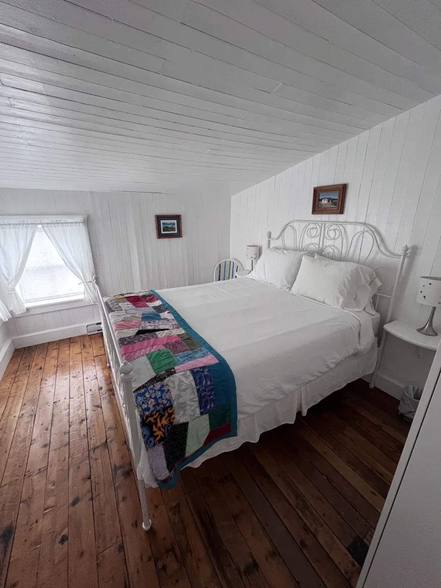 A white bedroom with wooden floors. It has a white metal framed bed with white bedding and a colourful quilt laid on it.