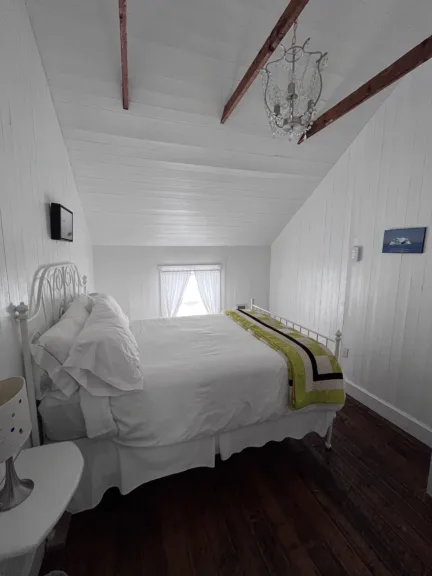 A white bedroom with wooden floors. It has a white metal framed bed with white bedding and a colourful quilt laid on it.