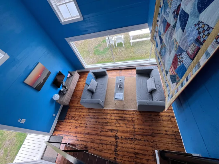 A top down view of a bright blue living room, with wooden floors and a large picture window on the back wall.