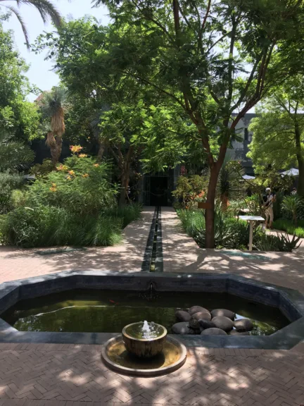 A small fountain is in front of a pond and has a path leading into some trees.