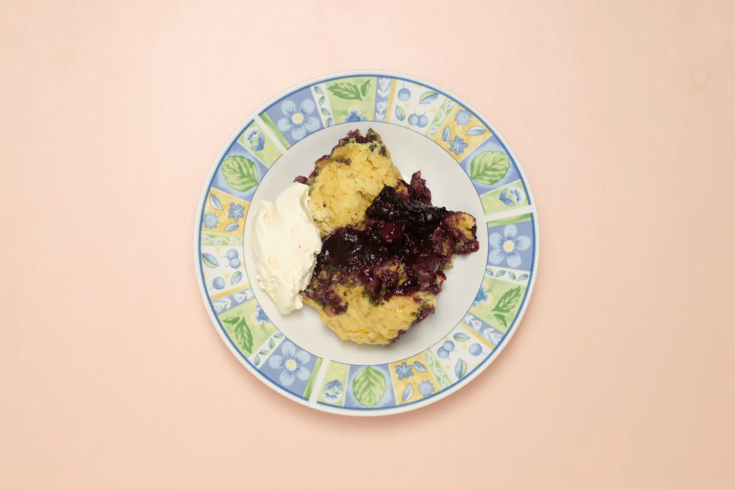 A white bowl with a floral pattern around the rim sits on a pink background. It contains blueberry grunt and ice cream.