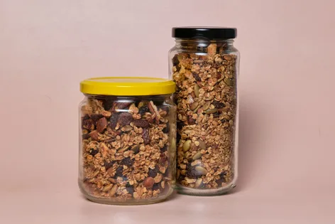 3 different sized glass jars of Maple Nut Granola on a pink background. One has a yellow lid, one a black lid and one a red lid.