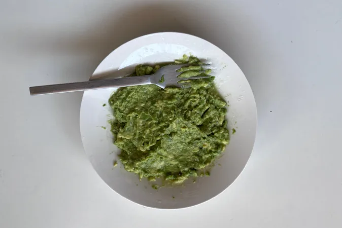 Mashed avocado in a white bowl, with a fork laid across it.