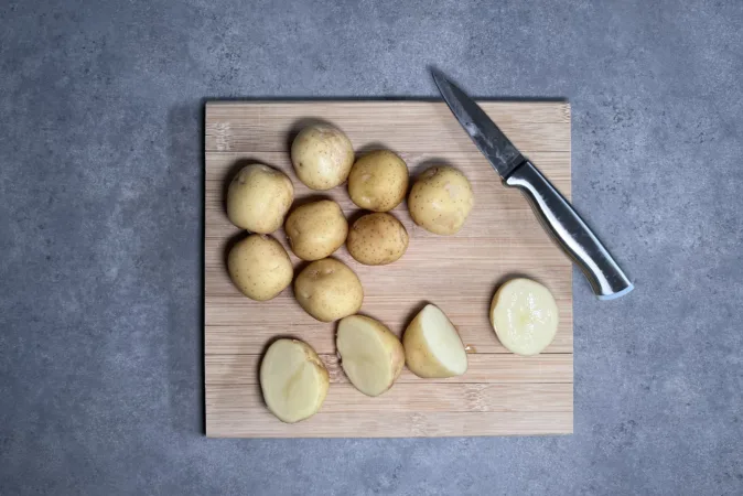 New potatoes and a small silver knife on a wooden chopping board. Two of the potatoes are cut in half.