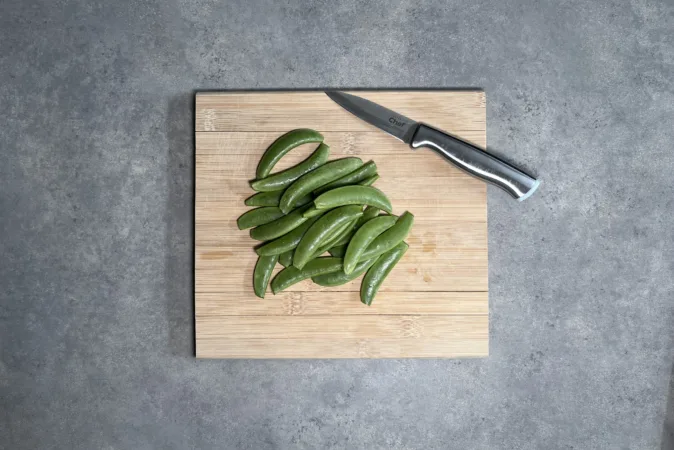 A pile of sugar snap peas and a small silver knife on a wooden chopping board.