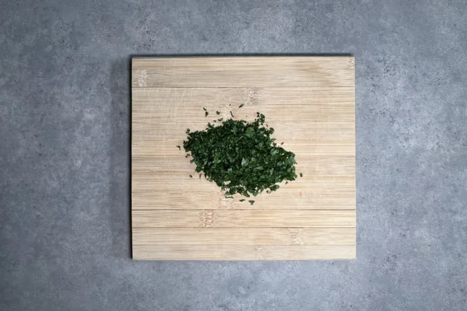 A small pile of chopped parsley on a wooden chopping board that is sat on a grey kitchen counter.