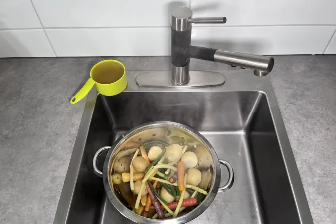 A colander full of colourful vegetables is draining in a metal sink. A green measuring cup, full of broth, sits next to the sink.