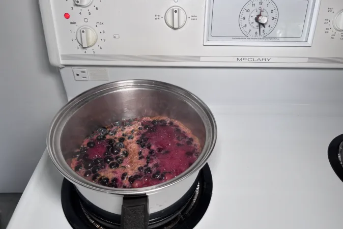 A metal saucepan is on a hob with blueberries simmering inside.