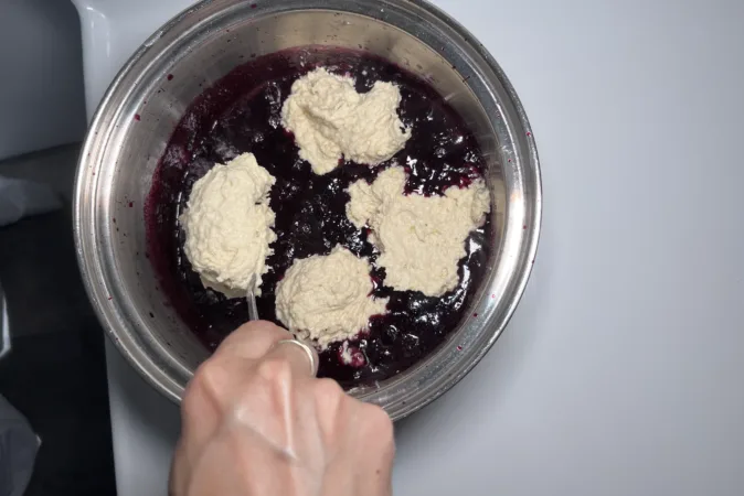 A metal saucepan with blueberries inside. A hand is spooning dumpling dough onto the fruit.